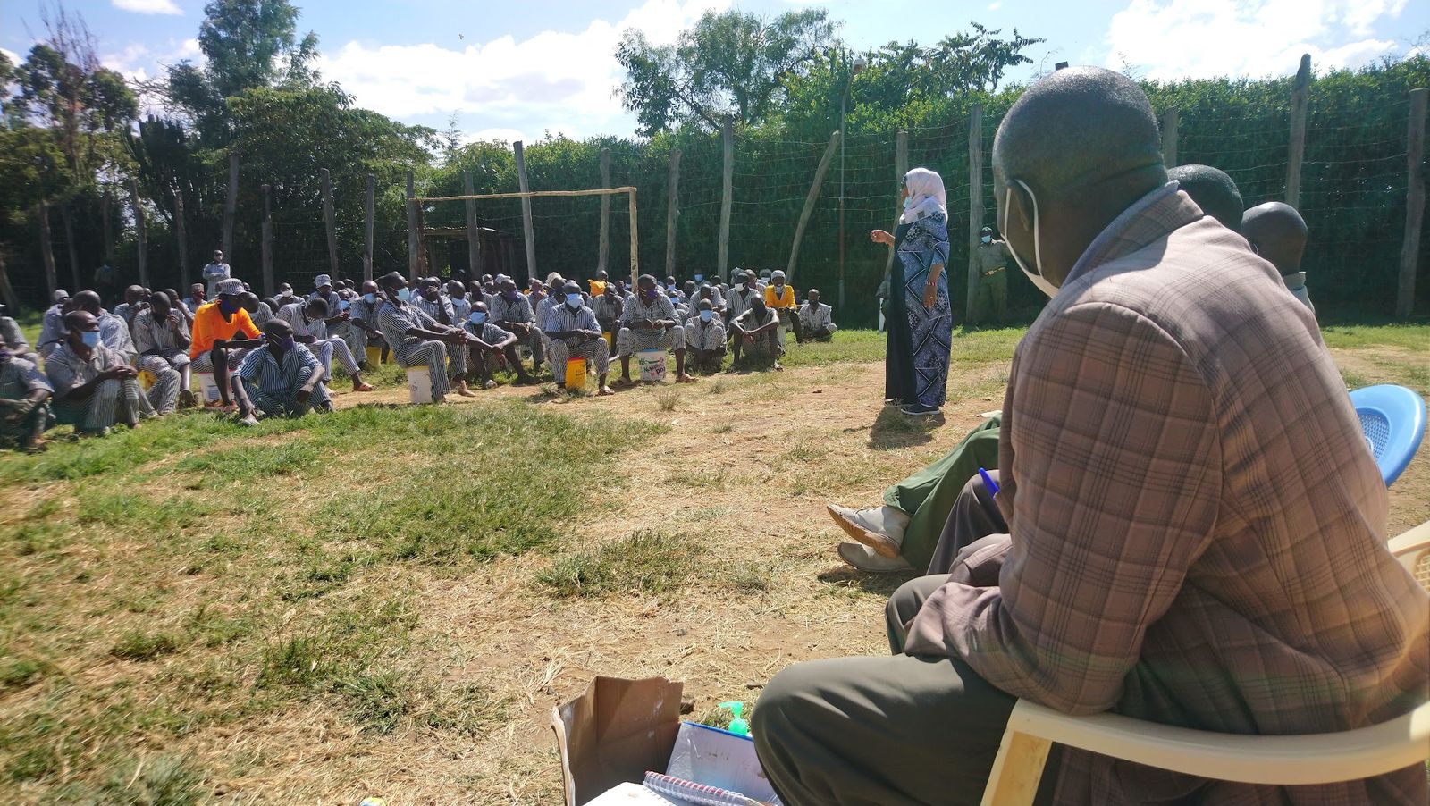 A facilitator addressing a large seated group of uniformed correctional officers and inmates during an MHPSS session